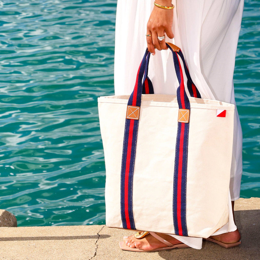 Person holding a white tote bag with red and blue straps by a body of water.
