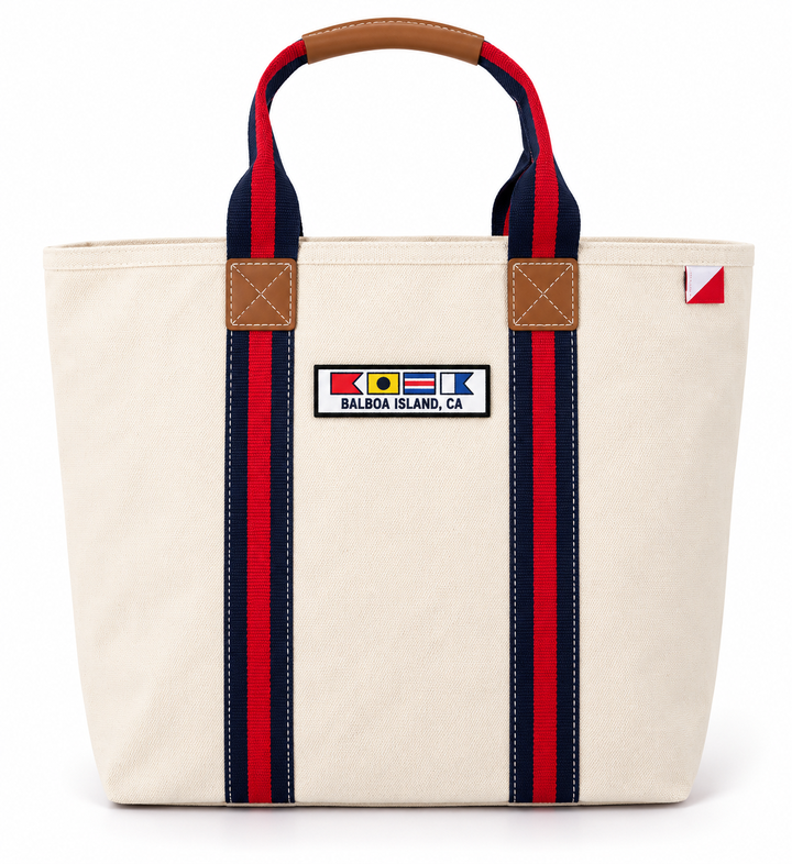 An off-white canvas tote bag with red and navy striped handles, brown leather accents, and a 'Balboa Island, CA' flag patch on the front.