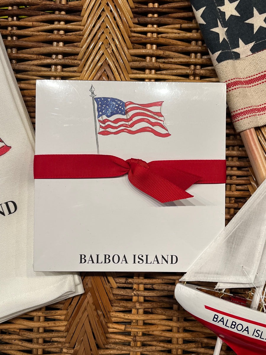 A white square gift box or card with a red ribbon bow across the middle, printed with an American flag and the words "BALBOA ISLAND," styled on a woven wicker surface with nautical props including a small model sailboat and a folded white towel.
