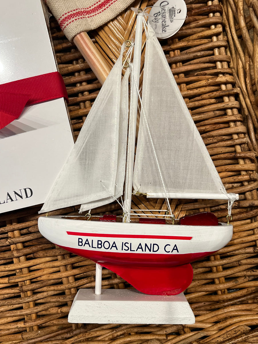 A decorative wooden sailboat ornament with white sails, a white hull, and a red lower section, printed with 'BALBOA ISLAND CA' on the side. It is displayed on a wicker tray next to a gift box.
