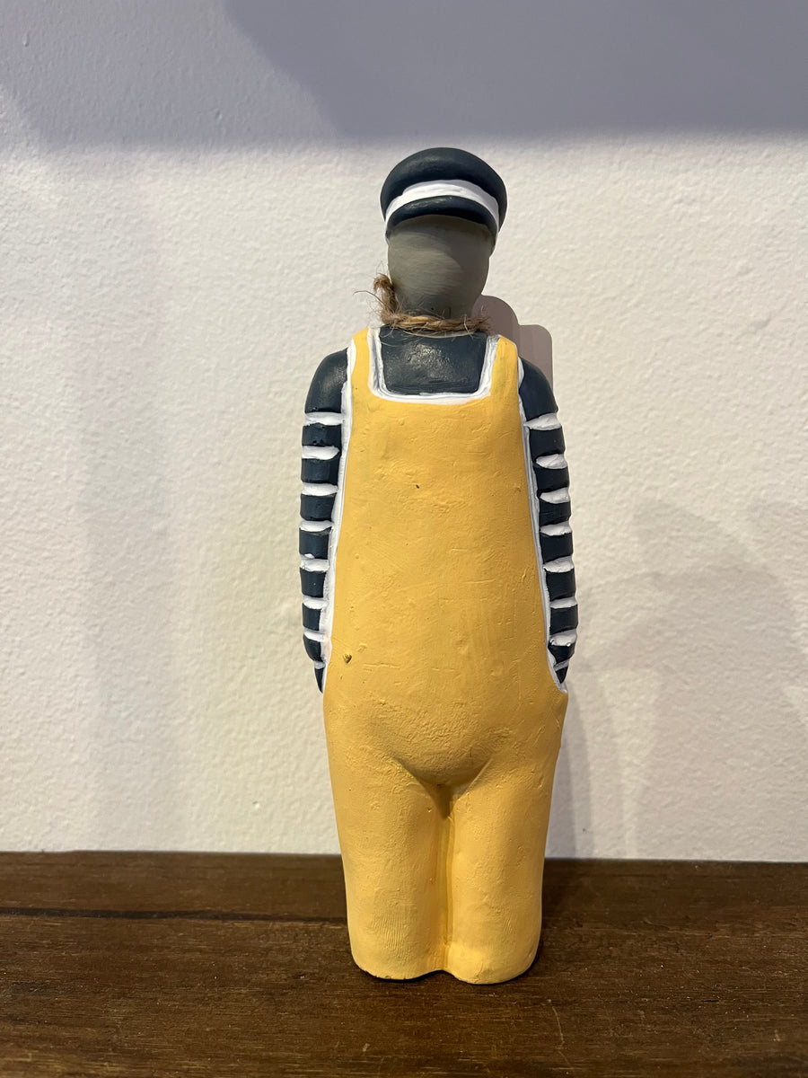 A hand-painted rustic wooden figurine of a sailor standing from the back, wearing a yellow overalls outfit, black and white striped shirt, and a striped hat, displayed on a wooden surface against a white wall.