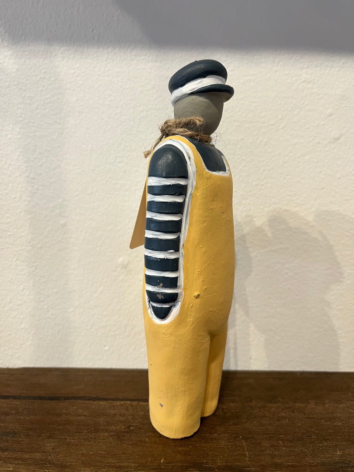 A hand-painted rustic figurine of a sailor or dockworker standing on a wooden surface against a textured off-white wall. The figure is painted with a mustard yellow body, a navy blue and white striped shirt, a navy blue and white striped hat, and a brown jute rope around the neck.