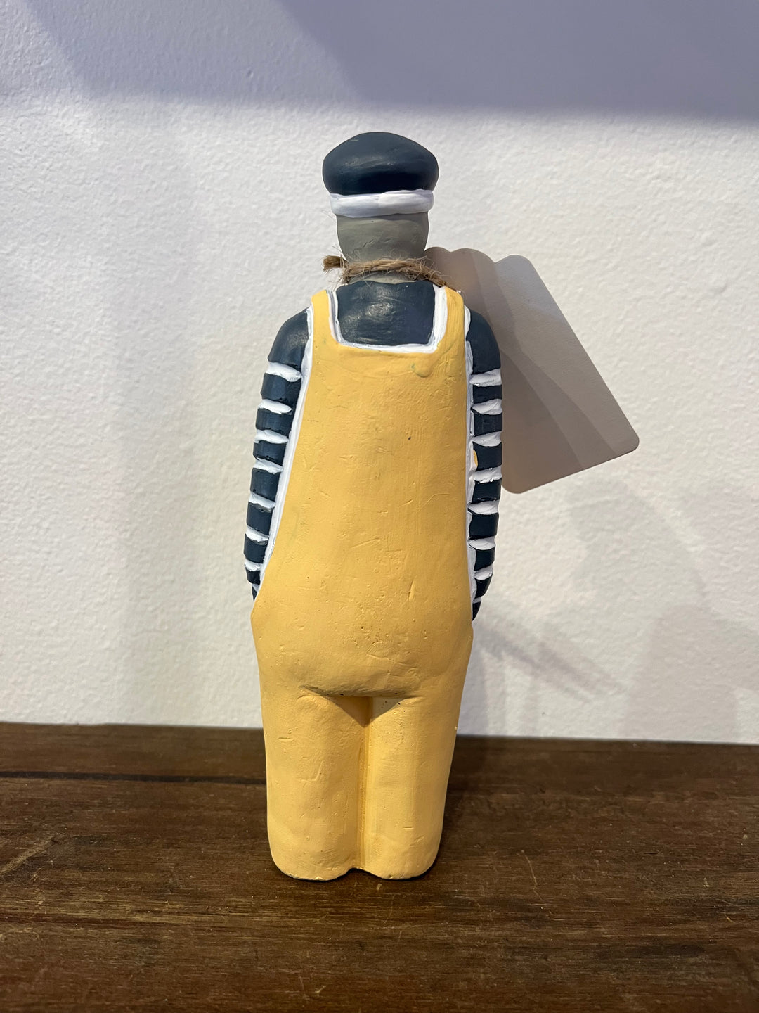 A hand-painted wooden figurine of a worker or sailor seen from the back, wearing a yellow overalls, a black and white striped shirt, and a black cap, standing on a wooden surface against a white wall.