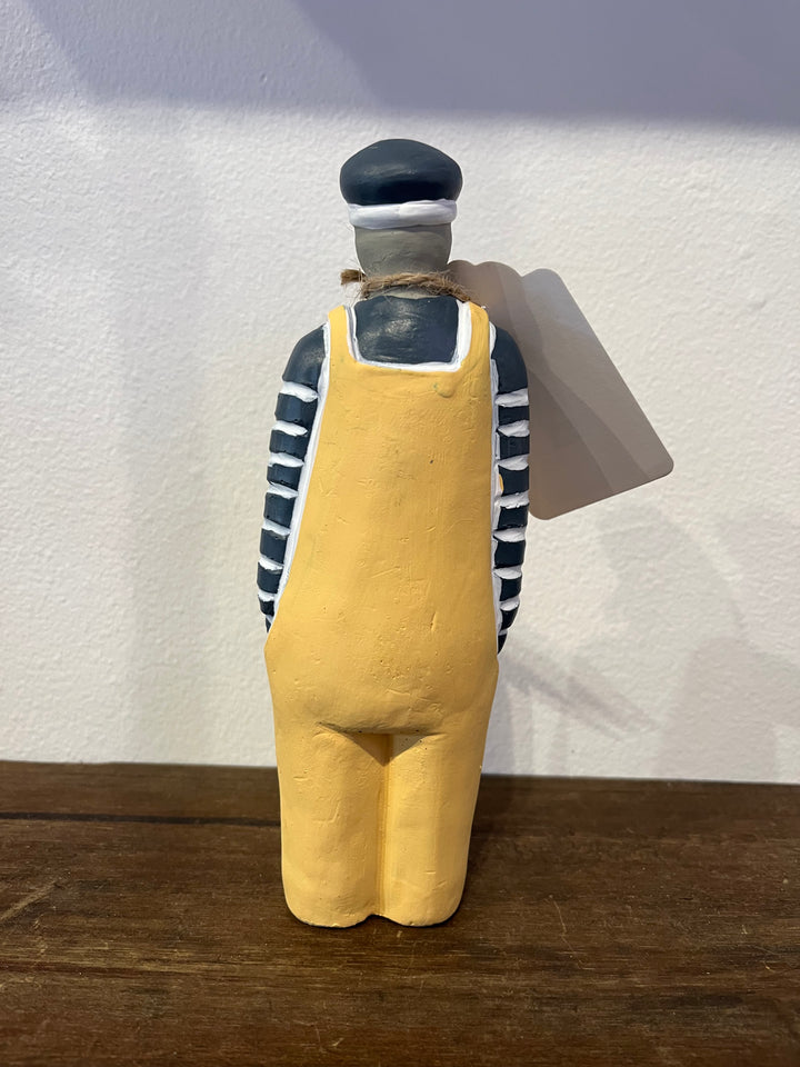 A hand-painted wooden figurine of a worker or sailor seen from the back, wearing a yellow overalls, a black and white striped shirt, and a black cap, standing on a wooden surface against a white wall.