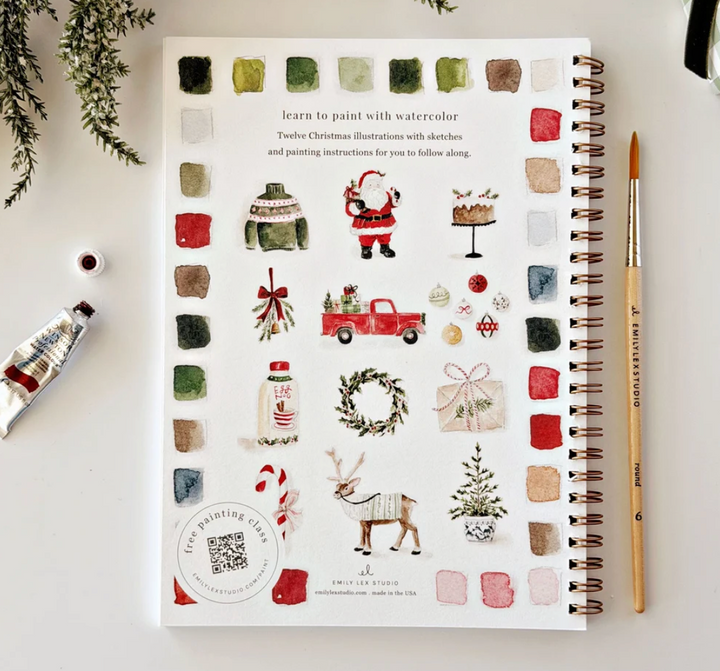 A spiral-bound watercolor painting book titled 'Teacher Christmas Illustration with sketches and painting instructions for you to follow along' by Emily Clark & Kathy Bouchard, shown on a white desk with a paint tube, paintbrush, and greenery.
