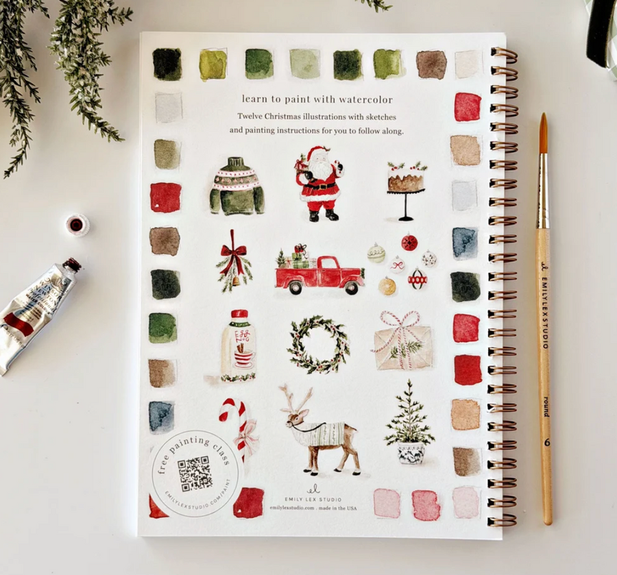 A spiral-bound watercolor painting book titled 'Teacher Christmas Illustration with sketches and painting instructions for you to follow along' by Emily Clark & Kathy Bouchard, shown on a white desk with a paint tube, paintbrush, and greenery.