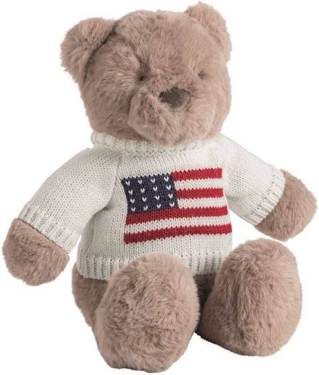 A plush teddy bear in light brown fur wearing a cream knit sweater with a red, white, and blue American flag design on the chest, shown against a plain white background.