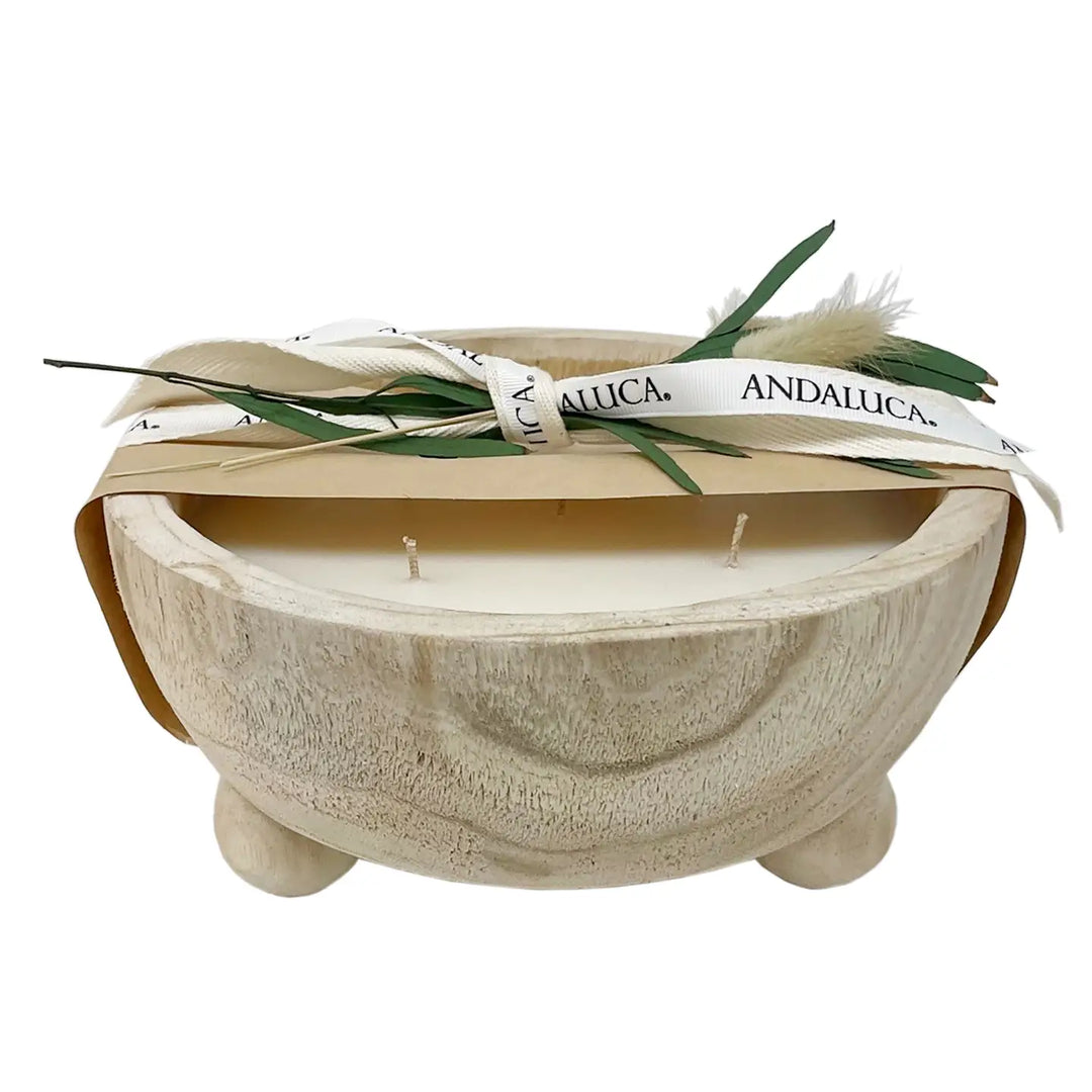 A three-wick soy candle in a carved light wood bowl, wrapped with a white ribbon printed with the brand name 'ANDALUCA' and decorated with green leaves and pampas grass, on a white background.