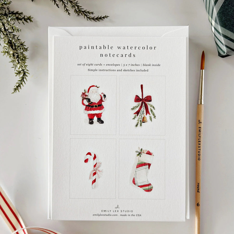 A flat-lay of a notepad card set titled 'paintable watercolor notecards' by Emily Lee Studio, featuring four sample watercolor illustrations of Santa Claus, mistletoe, a candy cane, and a Christmas stocking. The card is styled on a white surface with a paintbrush, evergreen branches, and striped fabric.