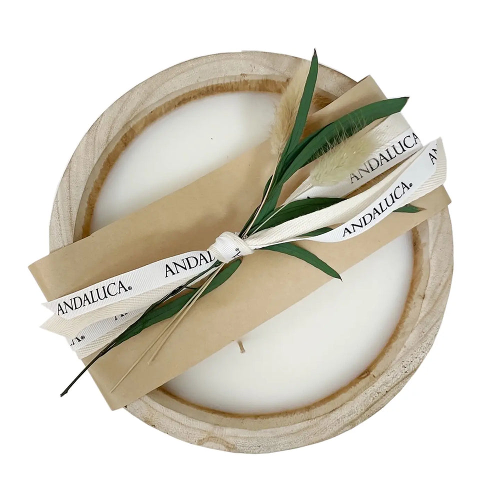 A round wooden candle holder containing white wax, wrapped with a kraft paper band and a white ribbon printed with the brand name 'ANDALUCÁ'. Green leaves and a dried feather are tucked into the wrap, all photographed on a clean white background.