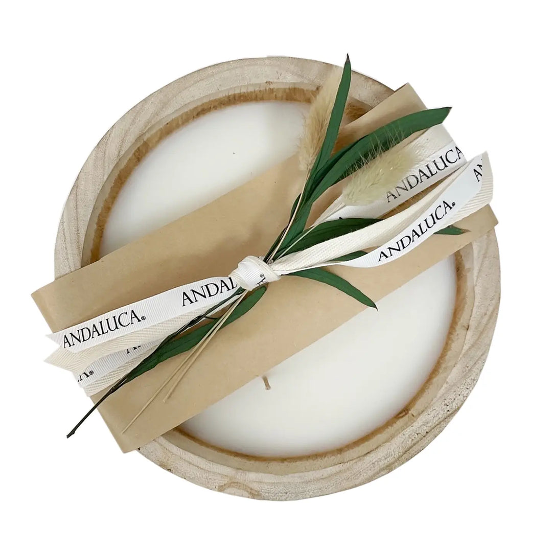 A round wooden candle holder containing white wax, wrapped with a kraft paper band and a white ribbon printed with the brand name 'ANDALUCÁ'. Green leaves and a dried feather are tucked into the wrap, all photographed on a clean white background.