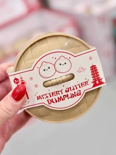A hand with red nail polish holds a round beige container labeled 'MYSTERY GLITTER DUMpling' with cartoon dumpling faces and red decorative graphics on a blurred pink-and-white background.