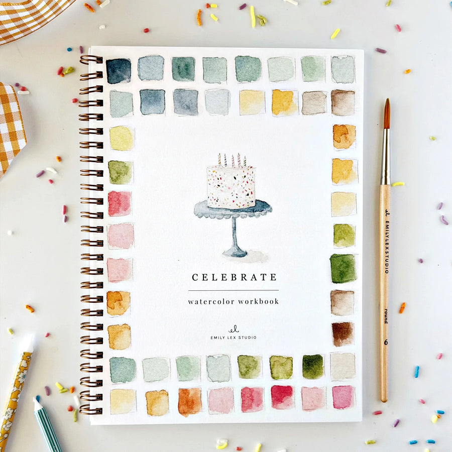 A spiral-bound watercolor workbook titled 'CELEBRATE watercolor workbook' by Emily Lee Studio, displayed on a light surface with colorful paint swatches around the border and a cake illustration in the center. A paintbrush and colored pencils are placed nearby.